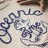 Close-up of a shirt with blue embroidered text and graphics on a beige background.